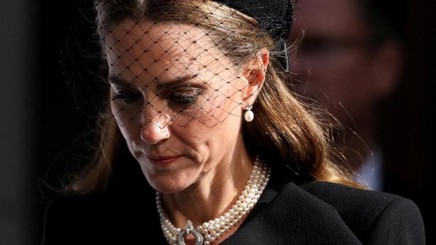 Prinzessin Kate, mit einem Schleierhut und einer Perlenkette, blickt gesenkt.