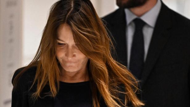 Carla Bruni blickt nach unten, im Hintergrund steht ein Mann im Anzug.