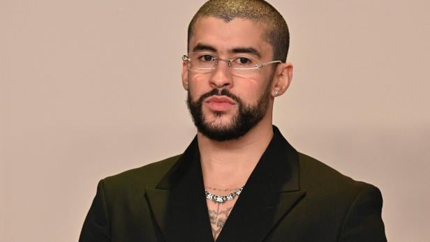Bad Bunny mit Brille, Bart und kurzem Haar trägt ein schwarzes Jackett und eine Halskette.