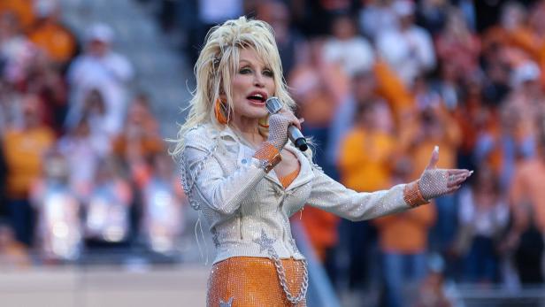 Dolly Parton singt in glitzernder Jacke und orangefarbener Hose vor einer Menschenmenge.