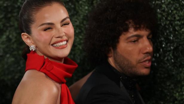 Selena Gomez lächelt in einem roten Kleid steht neben Benny Blanco im Anzug.