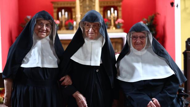 Drei Nonnen in Tracht stehen vor einem roten Altarraum.