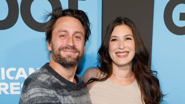 Kieran Culkin im Streifenpullover umarmt Jazz Charton in einem beigen Kleid. Er berührt ihren Babybauch.