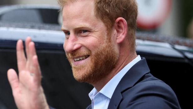 Prinz Harry lächelt und winkt in einem dunklen Anzug mit hellblauem Hemd.