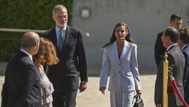 König Felipe und Königin Letizia gehen, umgeben von anderen Personen, in Anzügen.