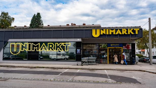 Ein Supermarkt mit dem Schriftzug „Unimarkt“ und mehreren Einkaufswagen vor dem Eingang, zwei Personen gehen hinein.