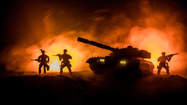 Drei bewaffnete Soldaten und ein Panzer stehen vor einem brennenden, rauchigen Hintergrund.