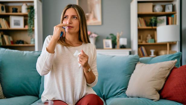Eine Frau sitzt auf einem Sofa und benutzt Nasenspray, während sie ein Taschentuch in der Hand hält.