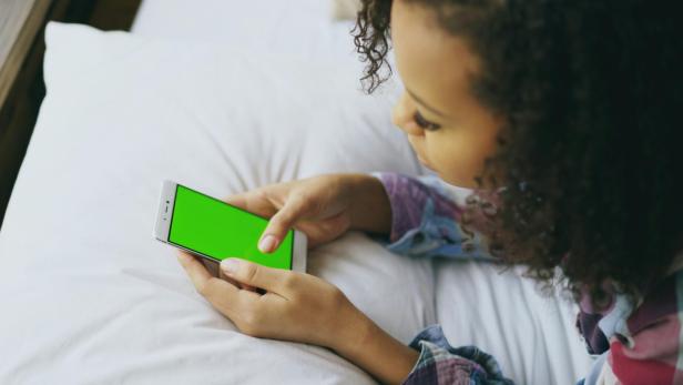 Jugendliche liegt im Bett und benutzt ein Smartphone mit grünem Bildschirm.