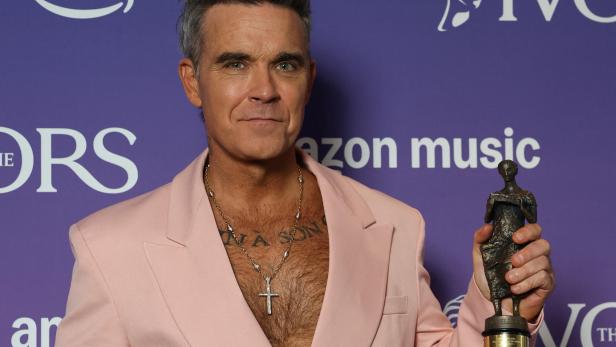 Robbie Williams im rosa Sakko hält eine Auszeichnung vor einem lila Hintergrund mit Amazon Music Logos.