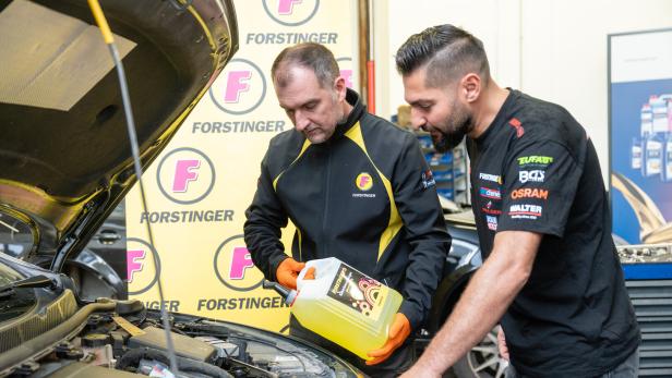 Zwei Männer prüfen in einer Autowerkstatt den Motorraum eines Fahrzeugs und füllen gelbe Flüssigkeit ein.