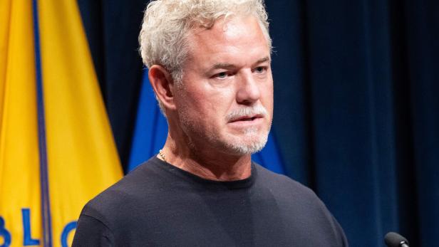 Eric Dane mit grauem Haar und Bart steht vor Flaggen und spricht in ein Mikrofon.