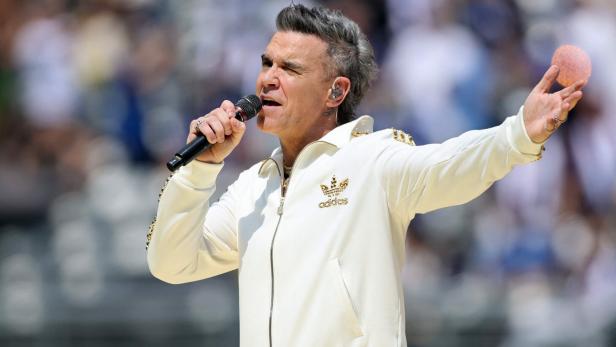 Robbie Williams singt mit Mikrofon in weißer Adidas-Jacke vor verschwommenem Hintergrund.
