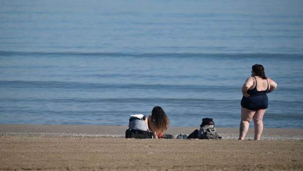 Zwei Frauen mit Taschen am Strand, eine sitzt im Sand, die andere steht im Badeanzug und blickt aufs Meer.