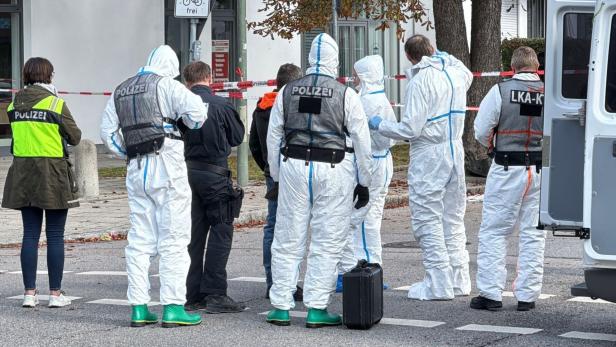 Mehrere Ermittler in Schutzanzügen und Polizeiwesten stehen an einem abgesperrten Tatort auf einer Straße.