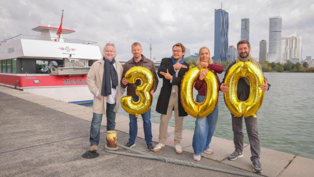 Fünf Personen stehen am Ufer mit goldenen Luftballons in Form der Zahl 300, im Hintergrund ein Schiff und moderne Hochhäuser.