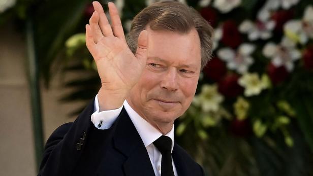 Großherzog Henri im dunklen Anzug mit Krawatte winkt vor einem floralen Hintergrund.