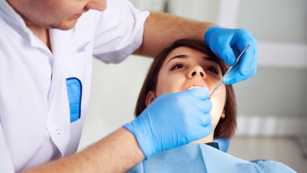 Eine Frau sitzt auf einem Behandlungsstuhl, während ein Zahnarzt mit blauen Handschuhen ihre Zähne untersucht.