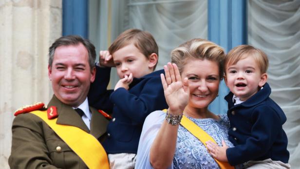 Großherzog Guillaume in Uniform und seine Frau winken von einem Balkon mit roter Balustrade.