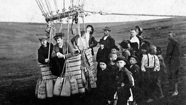 Eine schwarz-weiß-Aufnahme von mehreren Menschen rund um den und im Korb eines Heißluftballons, der am Boden befestig ist.