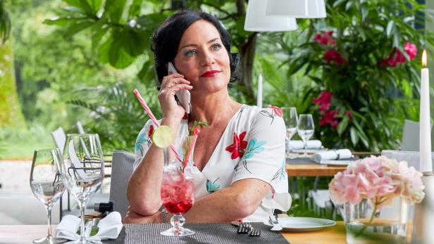 Eine Frau sitzt an einem gedeckten Tisch im Restaurant, telefoniert und hat einen Cocktail mit Limettenscheibe vor sich.