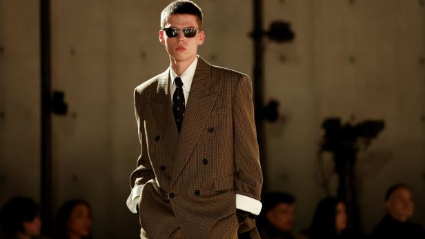 Juger Mann mit Zweireiher und Sonnenbrille am Laufsteg. Die Hände hat er in der Hosentasche.