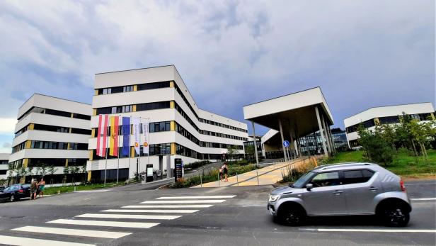 Mehrstöckiges modernes Gebäude mit mehreren Flaggen davor, Zebrastreifen und vorbeifahrendem silbernen Auto.