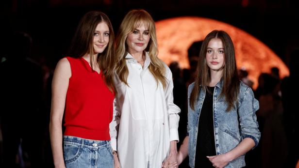 Nicole Kidman steht mit ihren Töchter Sunday und Faith Händchen haltend vor einem dunklen Hintergrund.