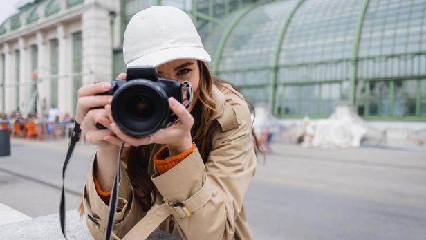 Eine Frau mit beiger Jacke und weißer Kappe hält eine Kamera und fotografiert vor dem Palmenhaus im Burggarten in Wien.