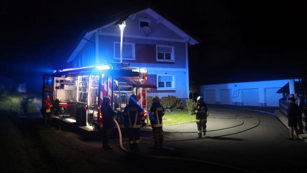 Ein Feuerwehrauto mit Blaulicht steht nachts vor einem Haus, Feuerwehrleute sind im Einsatz.