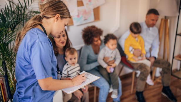 Eine Krankenschwester macht sich Notizen, während Familien mit Kindern im Wartezimmer sitzen.