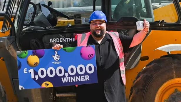 Mann (39) vor Baufahrzeug mit Lottogewinn in der Hand.