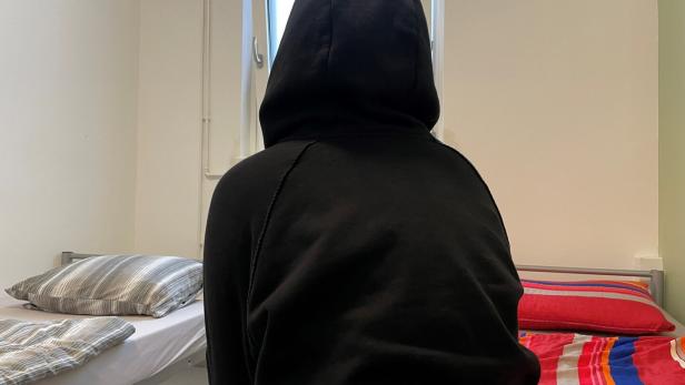 Junge Frau sitzt mit Kapuze auf dem Kopf verkehrt zur Kamera auf einem Bett