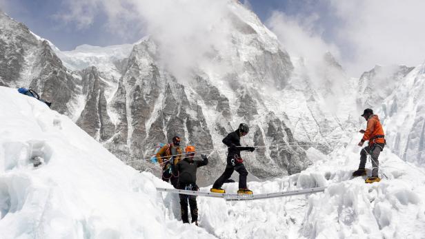 Vier Bergsteiger überqueren eine schmale Leiter über einer Gletscherspalte in verschneiter Hochgebirgslandschaft.
