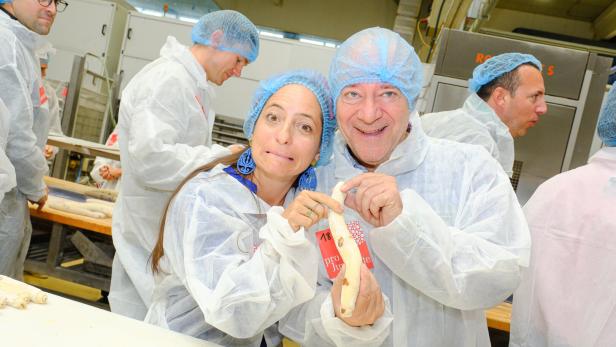 Caroline Athanasiadis und Andy Lee Lang in Schutzkleidung und Hauben halten lächelnd ein Teigstück in der Hand