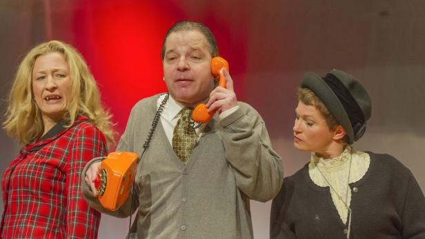 Drei Schauspieler auf einer Bühne, einer telefoniert mit einem orangefarbenen Telefon.