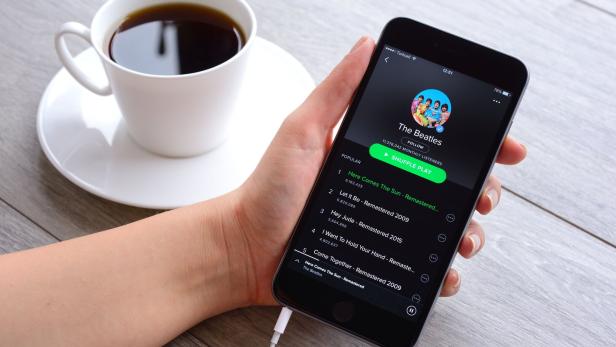 Eine Hand hält ein Smartphone mit geöffneter Musik-App, daneben steht eine Tasse schwarzer Kaffee auf einem Unterteller.