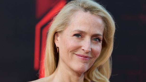 Gillian Anderson mit langen blonden Haaren lächelt vor einem dunklen Hintergrund mit roten Elementen.