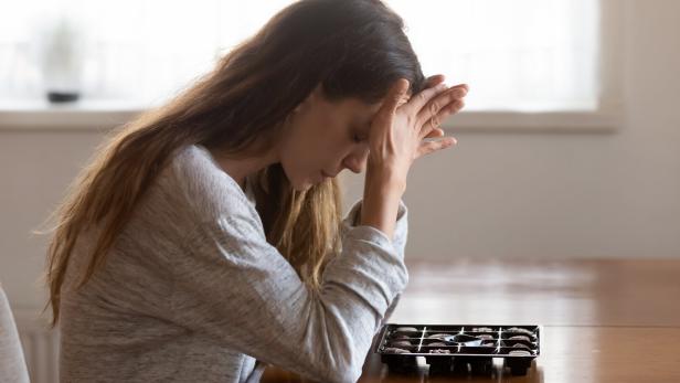 Menschen mit chronischen Schmerzen versuchen negative Gefühle oft über Essen zu regulieren, zeigt eine neue Untersuchung. Eine Frau sitzt mit gesenktem Kopf und verschränkten Händen vor einer Pralinenschachtel am Tisch.