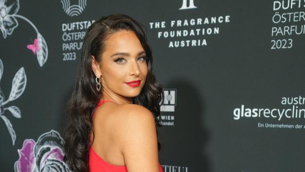 Amira Aly.jpg Amira Aly im roten Kleid blickt über die Schulter vor einem schwarzen Hintergrund mit Logos.