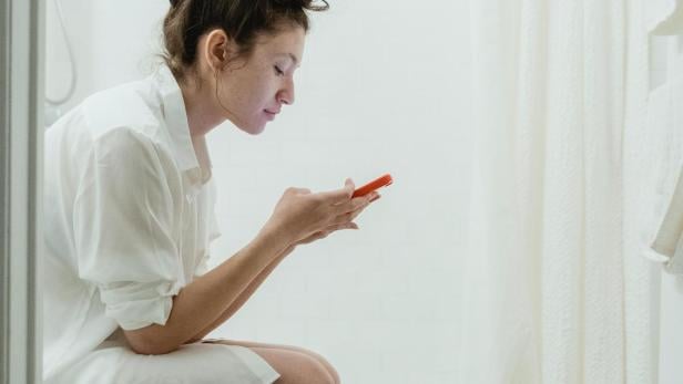 Nächtlicher Harndrang: Smartphone soll dahinterstecken. Frau sitzt mit Smartphone auf der Toilette.