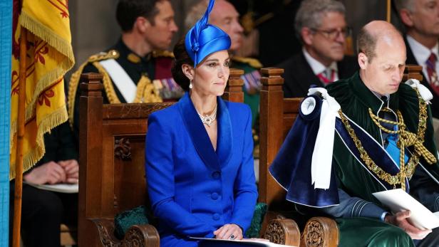 Prinzessin Kate.jpg Kate, in blauem Kostüm und Hut, sitzt neben William bei einer Veranstaltung.