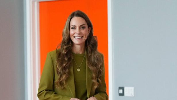 Prinzessin Kate trägt einen olivgrünen Hosenanzug und steht lächelnd in einem Raum mit orangefarbener Tür.
