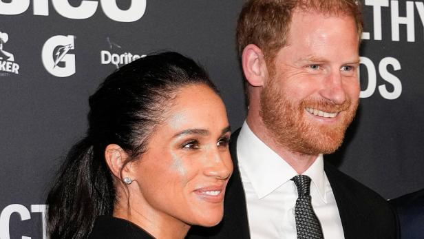 Meghan und Prinz Harry lächeln auf einer Veranstaltung vor einem dunklen Hintergrund.
