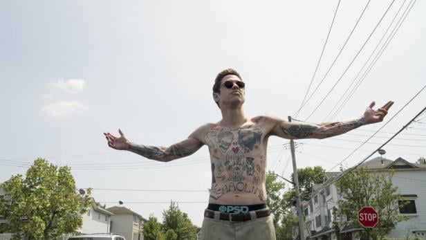 Szene aus dem Film The King of Staten Island (2020): Pete Davidson steht mit nacktem Oberkörper, der mit Tattoos bedeckt ist, und ausgebreiteten Armen und Sonnenbrille auf einer Straße in einem Wohngebiet.