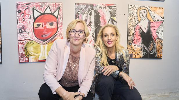 Zwei Frauen sitzen vor farbenfrohen, modernen Gemälden an einer Wand und lächeln in die Kamera.