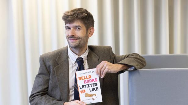Ein Mann im Anzug hält ein Buch mit dem Titel „Bella Barks Letztes Like“ und lächelt leicht.