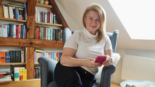 Eine blonde Frau sitzt mit ihrem pinken Handy in einem grauen Ikeastuhl vor einer Bücherwand.