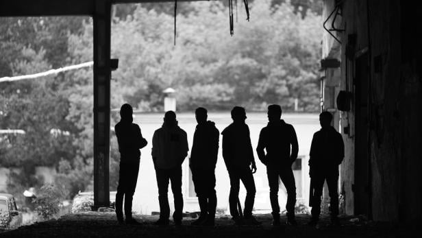 Sechs Personen stehen im Schatten eines Gebäudes und blicken nach draußen, im Hintergrund sind Bäume zu sehen.