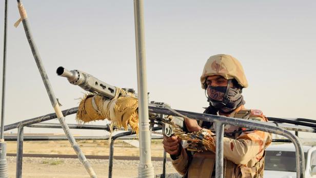 Ein Soldat in Tarnuniform und Helm steht hinter einem montierten Maschinengewehr und hält einen Munitionsgurt.